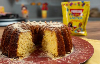 Fotografia de um bolo de milho com farinha láctea apoiado em um prato grande, raso e decorativo de tom vinho. Atrás do bolo tem uma embalagem de farinha láctea.