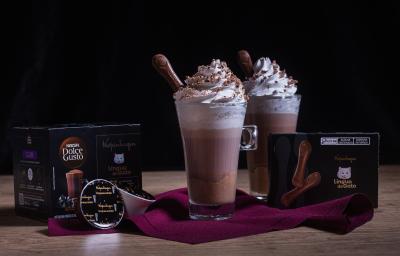 Fotografia em tons escuros de duas xícaras de vidro longas com Chococcino Mousse Língua de Gato, chantilly e língua de gato sobre um pano vinho. Ao lado tem uma caixa de nescafé e a outra de língua de gato. .