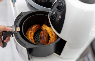 Fotografia dem tons claros de uma mão puxando a cestinha da air fryer branca com uma receita de banana douradinha.