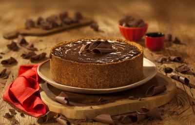 Fotografia em tons de marrom em uma bancada de madeira de cor marrom. Ao centro, uma tábua de madeira contendo o cheesecake e ao redor há pedaços de chocolates espalhados. Fotografia em tons de marrom em uma bancada de madeira de cor marrom. Ao centro, uma tábua de madeira contendo o cheesecake e ao redor há pedaços de chocolates espalhados.