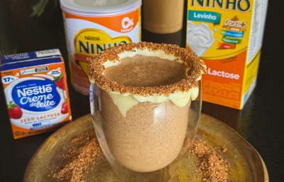 Fotografia de uma xícara de vidro com uma bebida de ninho com o creme de ninho na borda e ninho tostado também na borda e no prato raso em que a xícara está apoiada. O prato está sobre uma mesa preta, ao lado do prato tem produtos zero lactose nestlé